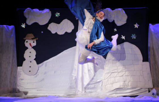 Scena spektaklu dla dzieci. Na pierwszym planie dziewczyna - śnieżynka w podskoku, za nią bałwan, zwały śniegu, obłoki na niebie.