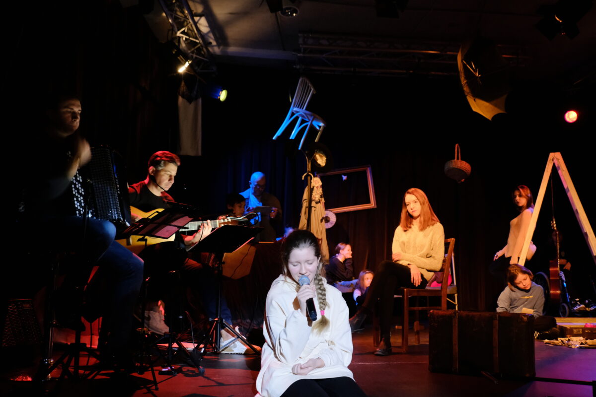Grupa młodzieży na scenie, siedzą w różnych miejscach, na środku stoi drabina, pod sufitem wiszą różne przedmioty, m. in. krzesło i parasol. Dziewczyna na pierwszym planie trzyma w dłoni mikrofon.