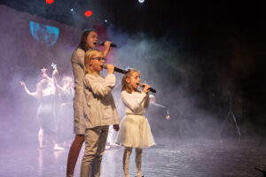 Dwie małe dziewczynki i nastolatka na scenie, wszystkie z mikrofonami w dłoniach