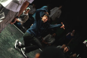 Nastoletni chłopak w pozie tanecznej na scenie, za nim na podłodze siedzi publiczność