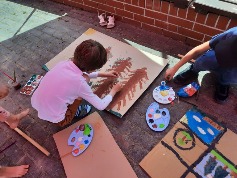 Chłopiec maluje farbami drzewa na dużym kartonie, zdjęcie z góry, nie widać twarzy chłopca