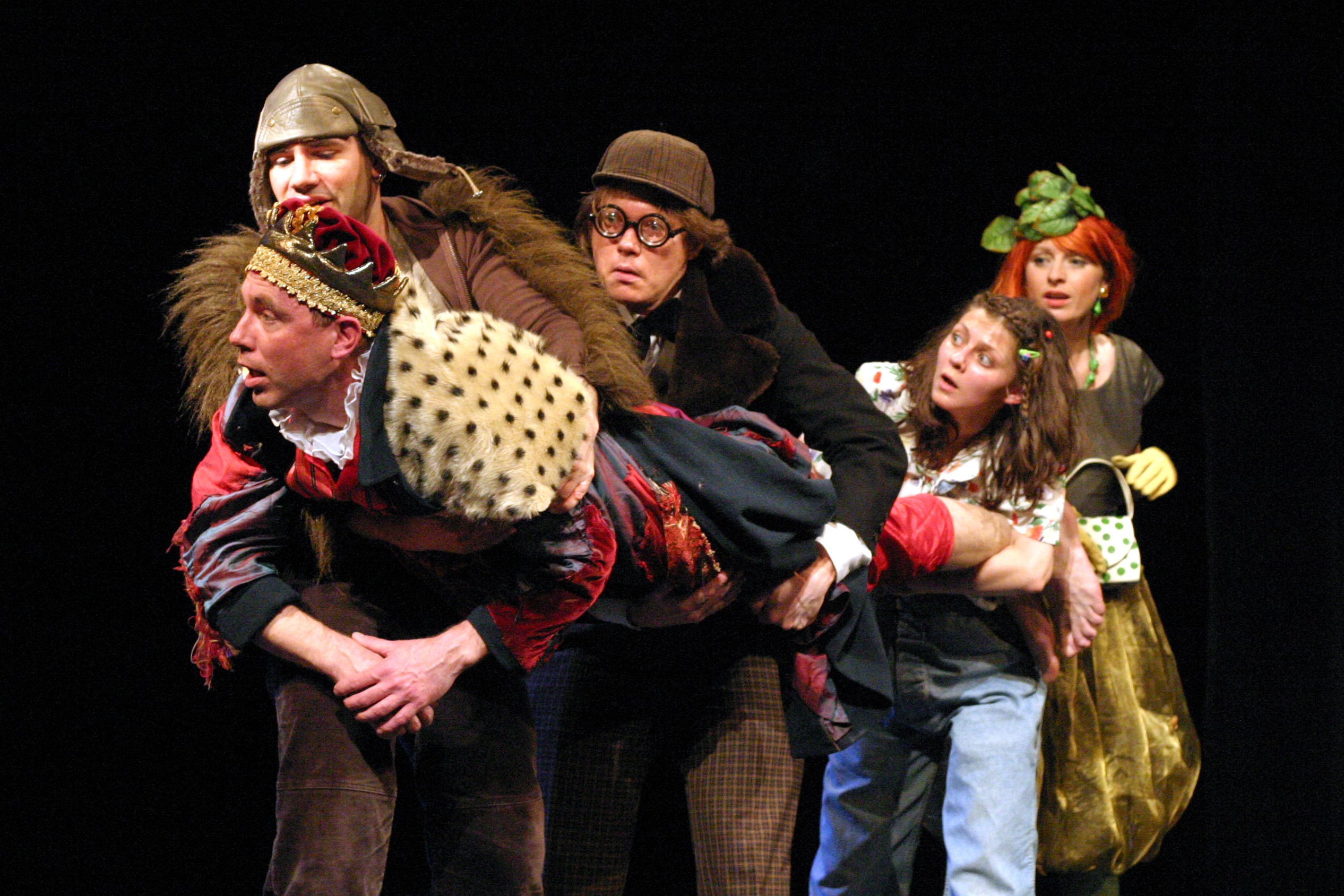 Pięć postaci na scenie w bajkowych kostiumach. Dwóch mężczyzn i dwie kobiety trzymają mężczyznę w kostiumie króla