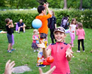 Grupa dzieci żonglujących piłeczkami na trawie, na pierwszym planie uśmiechnieta dziewczynka w czapce
