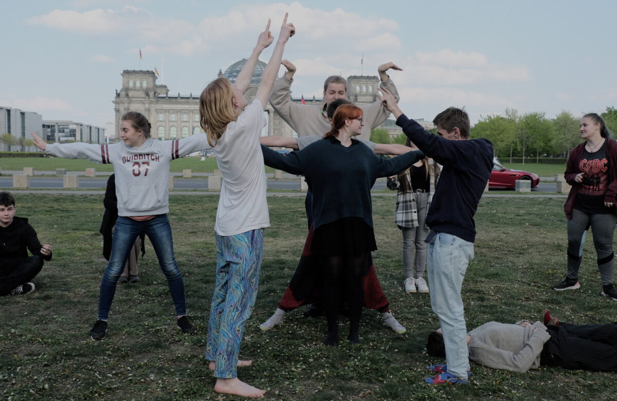 Grupa młodzieży na trawniku, każdy w innej pozie, jakby odgrywali jakąś scenkę. Za nimi duży budynek