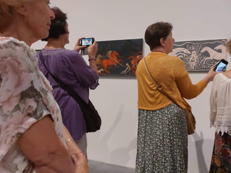 Grupa kobiet ogląda wystawę obrazów, niektóre robią zdjęcia telefonami komórkowymi