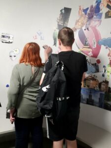 dwie osoby, kobieta i mężczyzna, stojący tyłem do obiektywu patrzą na białą ścianę udekorowaną magnetycznymi obrazkami