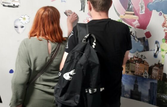 dwie osoby, kobieta i mężczyzna, stojący tyłem do obiektywu patrzą na białą ścianę udekorowaną magnetycznymi obrazkami