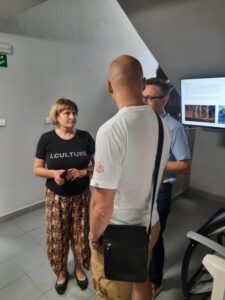 Trzy osoby, kobieta i dwóch mężczyzn, stoją na klatce schodowej i rozmawiają. W tle widać włączony ekran telewizora.