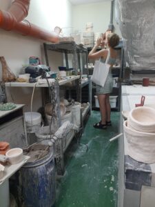 Kobieta w sukience i z torbą na ramieniu stoi między półkami zastawionymi pracami z gliny. W rękach trzyma telefon i robi zdjęcie jednemu z przedmiotów