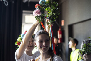 Na pierwszym planie uśmiechnięta dziewczyna, za nią druga próbuje założyć jej na głowę wianek z żywych kwiatów