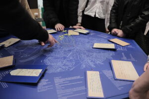 Zdjęcie przedstawia stół z historyczną mapą Warszawy i zapiskami na przymocowanych do niej kartkach. Widać postaci 4 osób, nie widzimy ich twarzy tyko dłonie pokazujące miejsca na mapie.