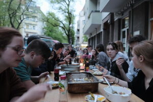 Zdjęcie przedstawia długi stół zastawiony jedzeniem. Wokół niego siedzą młode osoby, rozmawiają i jedzą.