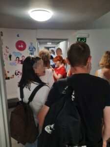 Grupa osób stoi tyłem do obiektywu i ogląda kolorowe zdjęcia przyklejone do białych ścian.