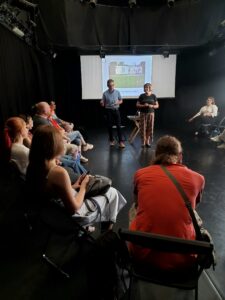 Zdjęcie przedstawia dużą czarną salę, na której w kręgu siedzi grupa około osób. Na środku stoi kobieta i mężczyzna mówiący do grupy. W tle widać ekran z prezentacją.