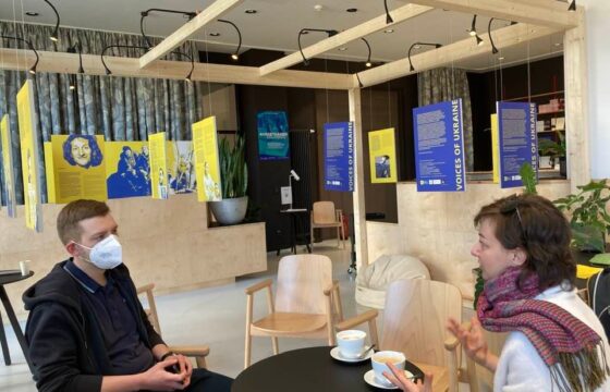 Dwie osoby siedzą przy stoliku w holu Instytutu Pileckiego i rozmawiają. W tle widać zawieszoną wystawę zdjęć z Ukrainy.