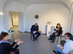 Cztery osoby siedzą w dużej białej sali w kręgu, dosyć oddalone od siebie. Kobieta po lewej stronie mówi gestykulując rękami.