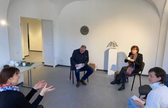 Cztery osoby siedzą w dużej białej sali w kręgu, dosyć oddalone od siebie. Kobieta po lewej stronie mówi gestykulując rękami.