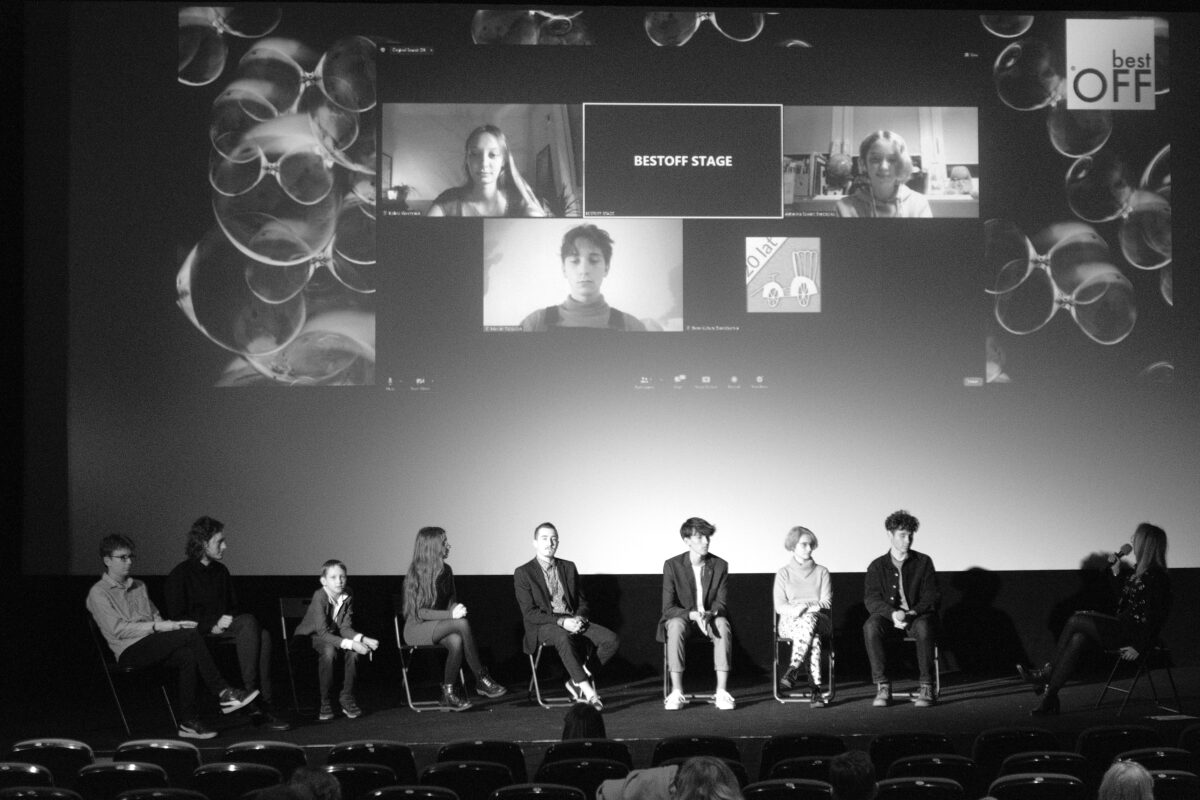 zdjęcie czarnobiałe, na scenie, na krzesłach siedzi dziewięć osób, chłopcy i dziewczynki, nad nimi na ekranie kinowym wyświetlają się 3 okienka rozmowy internetowej z trzema osobami