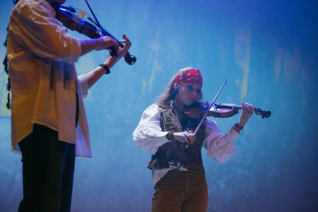 Dwie dziewczyny na scenie grają na skrzypcach, ta z prawej strony ucharakteryzowana jest na pirata