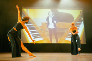 Dwie tancerki na scenie, za nimi na ekranie dwie klawiatury fortepianu, między nimi stoi kobieta w średnim wieku