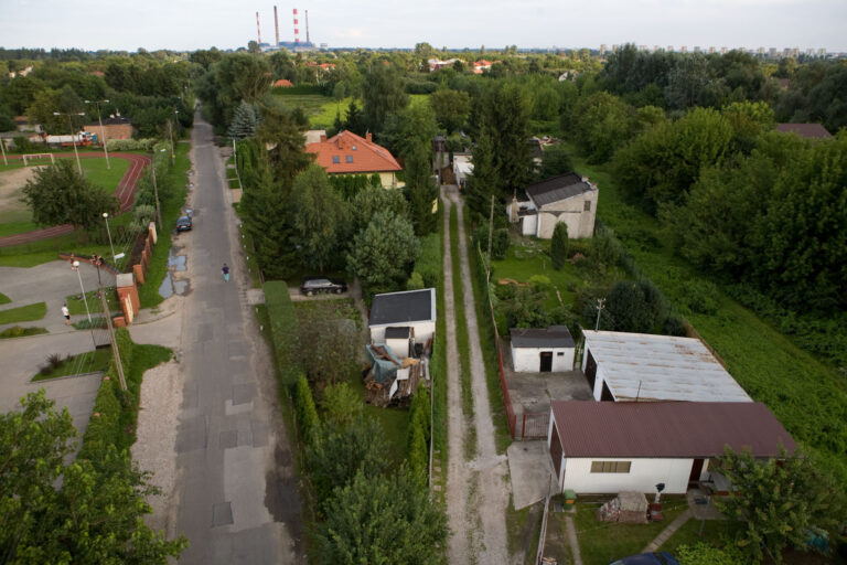 Wąska asfaltowa droga, po jej lewej stronie boisko, po prawej budynki. Po obu stronach drogi drzewa. Zdjęcie zrobione z góry