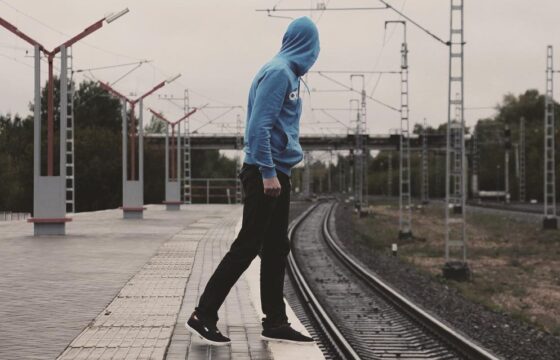 Mężczyzna w bluzie, z kapturem na głowie stoi na brzegu peronu przy torach