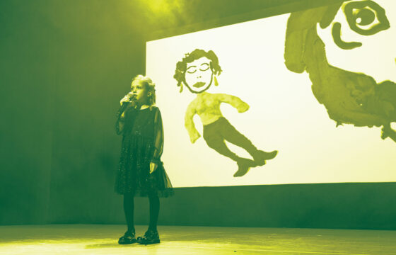 Dziewczynka z mikrofonem w dłoni na scenie, za nią na ekranie rysunek postaci