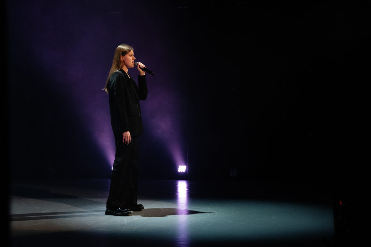 Nastolatka, ubrana w koszulę i spodnie, ma długie włosy, stoi na scenie z mikrofonem w dłoni.