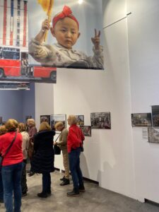 Grupa kobiet zwiedza wystawę fotograficzną, zdjęcia zawieszone są na ścianach, nad głowami wisi duże zdjęcie skośnookiego dziecka.