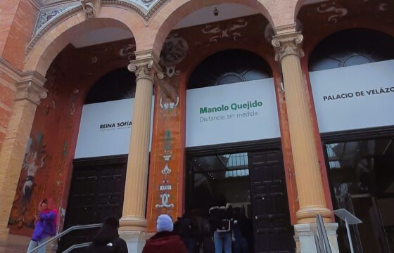 Dwie osoby wchodzą po schodach do dużego budynku z arkadami. Nad drzwiami widnieje napis z nazwiskiem malarza Manolo Quejido. Jest to galeria Muzeum Sztuki Współczesnej Królowej Zofii.