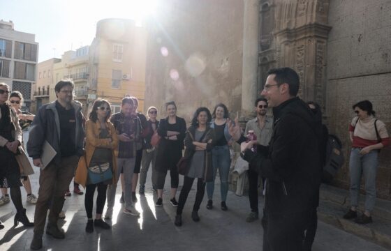 Grupa około 20 osób stoi wokół mówiącego i gestykulującego mężczyzny w okularach. Zdjęcie przedstawia grupę na ulicy, w słoneczny dzień.