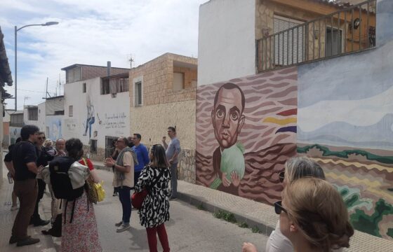 Wąska ulica z niskim zabudowaniem. Ściany budynków mają namalowane murale. Grupa około 20 osób stoi na ulicy i słucha jednego mężczyzny.