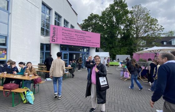 Młoda uśmiechnięta kobieta z torbą na ramieniu z dużym logotypem Dorożkarnia stoi przed budynkiem. Przy budynku stoły, przy których siedzą młodzi ludzie, obok niej przechodnie.