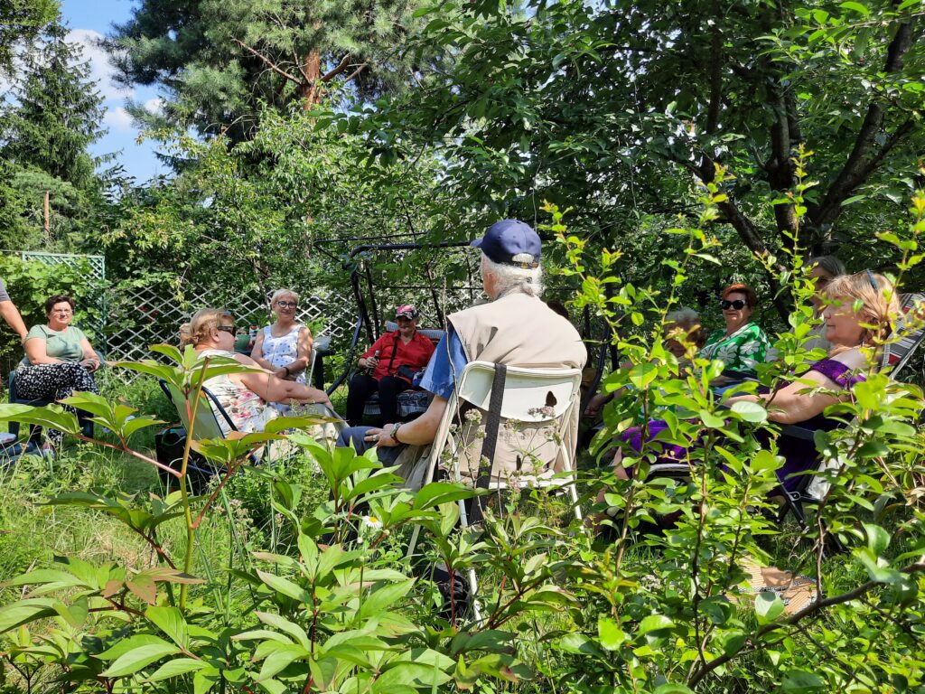 Grupa seniorek i seniorów siedzi w kręgu w ogrodzie, jest lato