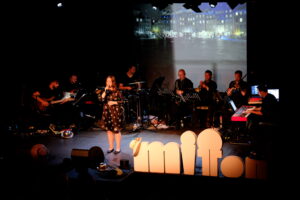 Dziewczyna z mikrofonem w dłoni na scenie, za nią zespół muzyczny, przed nią napis MIT TON