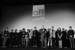 Grupa młodych osób na scenie, stoją pod napisem Best Off Przegląd Najlepszych Filmó Polskiego Kina Młodych 2022, przed nimi starszy mężczyzna z mikrofonem w dłoni
