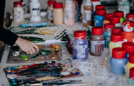 Stół zastawiony farbami akrylowymi oraz dłonie osoby malującej obraz