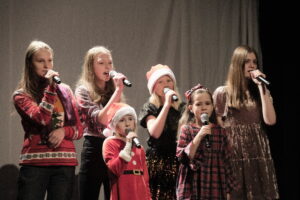 Grupa dziewcząt w róznym wieku na scenie, z mikrofonami w dłoniach, dwie z nich na głowach mają czapki Mikołaja.