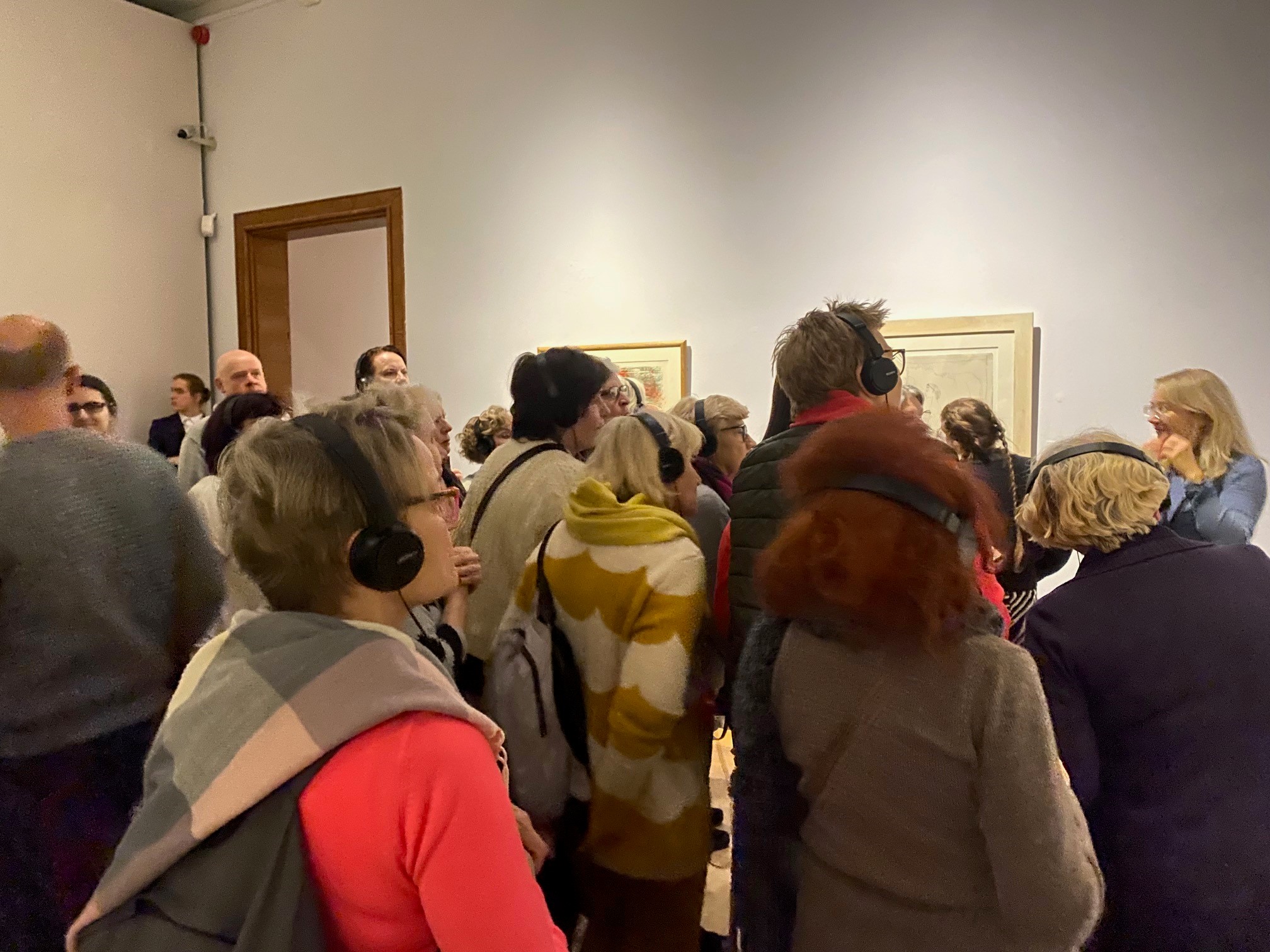 Grupa osób zgromadzona w pomieszczeniu wystawowym, nad ich głowami fragmenty obrazów zawieszonych na ścianie