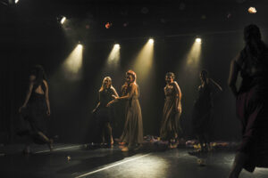 kilka kobiet na scenie w długich sukienkach, stoją oświetlane reflektorami z góry