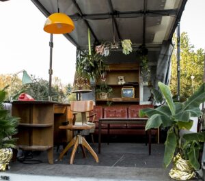 Zaaranżowany pokój na pace samochodu dostawczego, jest kanapa, biurko, krzesło, kwiaty doniczkowe, nad biurkiem wisi lampa.