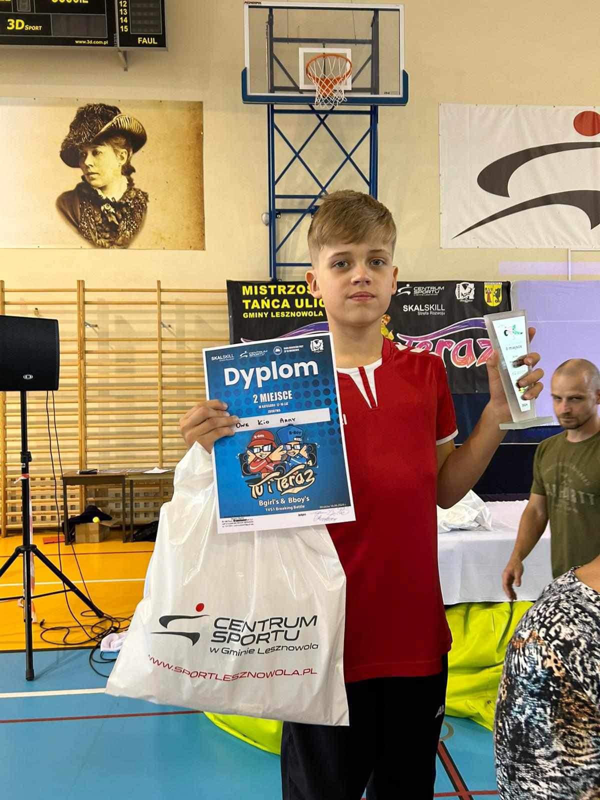 Uśmiechnięty chłopiec z dyplomem i reklamówką w jednej ręce oraz statuetką w drugiej.