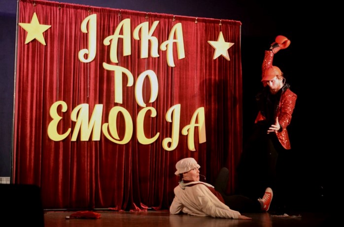 Scena, na scenie dwie osoby, jedna leży, druga nad nią z ręką w górze. Za postaciami kurtyna z napisem "Jaka to emocja?"