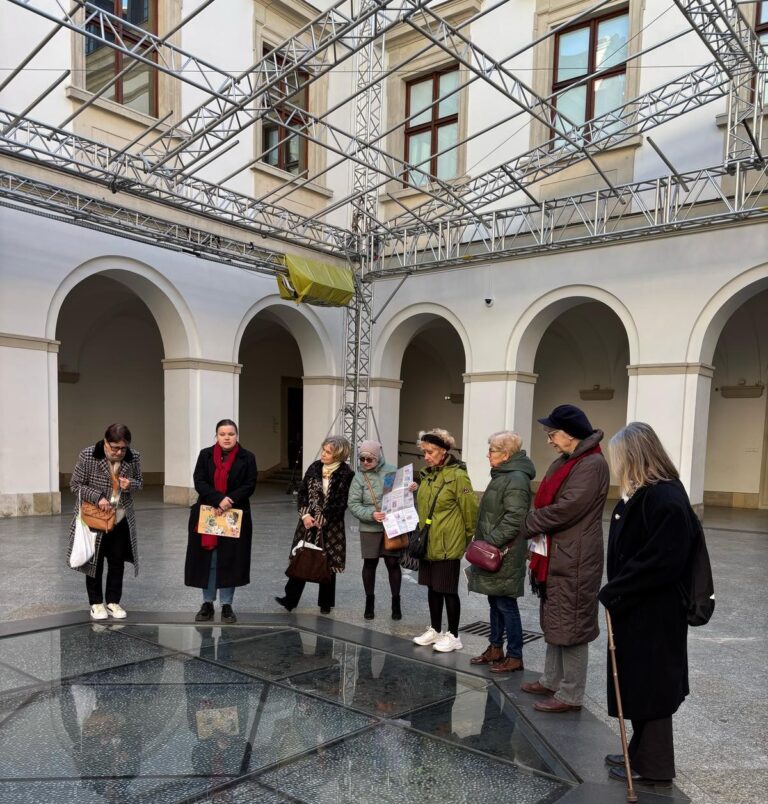 Grupa ośmiu osób stoi wpatrzona w coś na podłodze lub chodniku. Wszyscy ubrani są w stroje zimowe, za nimi budynek.
