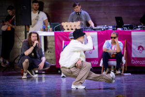 Chłopak w kapeluszu na scenie, prawdopodobnie tańczy breakdance, za nim siedzi i stoli kilka osób