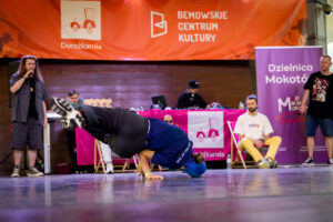 Osoba na scenie, tańczy breakdance, jest w pozycji na podpartych rękach. Za nim kilka osób siedzą i stoją osoby. Na d tancerzem wisi baner z logotypami Dorożkarni i Bemowskiego Centrum Kultury