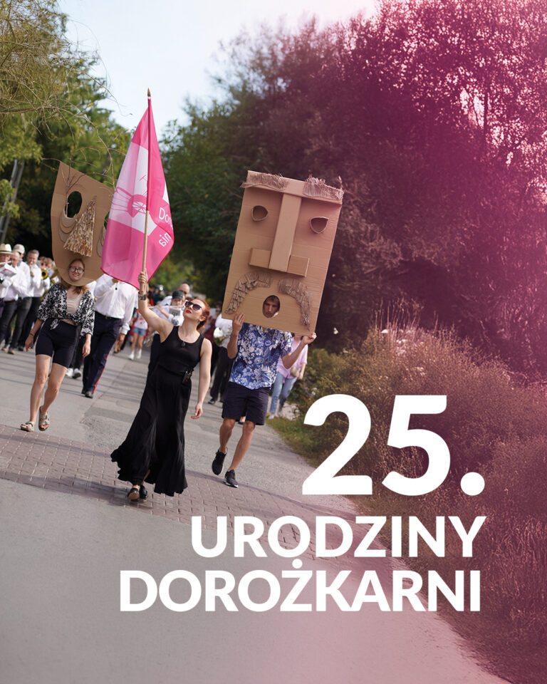 Grupa osób idzie brukowaną drogą wśród drzew i krzewów, osoby z przodu niosą flagę i dekorację z tektury