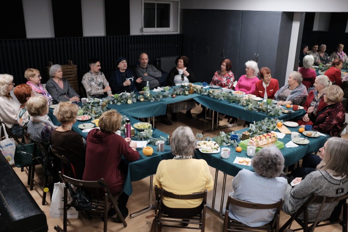 Fotografia przedstawiająca osoby seniorskie siedzące przy wspólnym, kwadratowym stole. Stół jest wypełniony naczyniami z jedzeniem